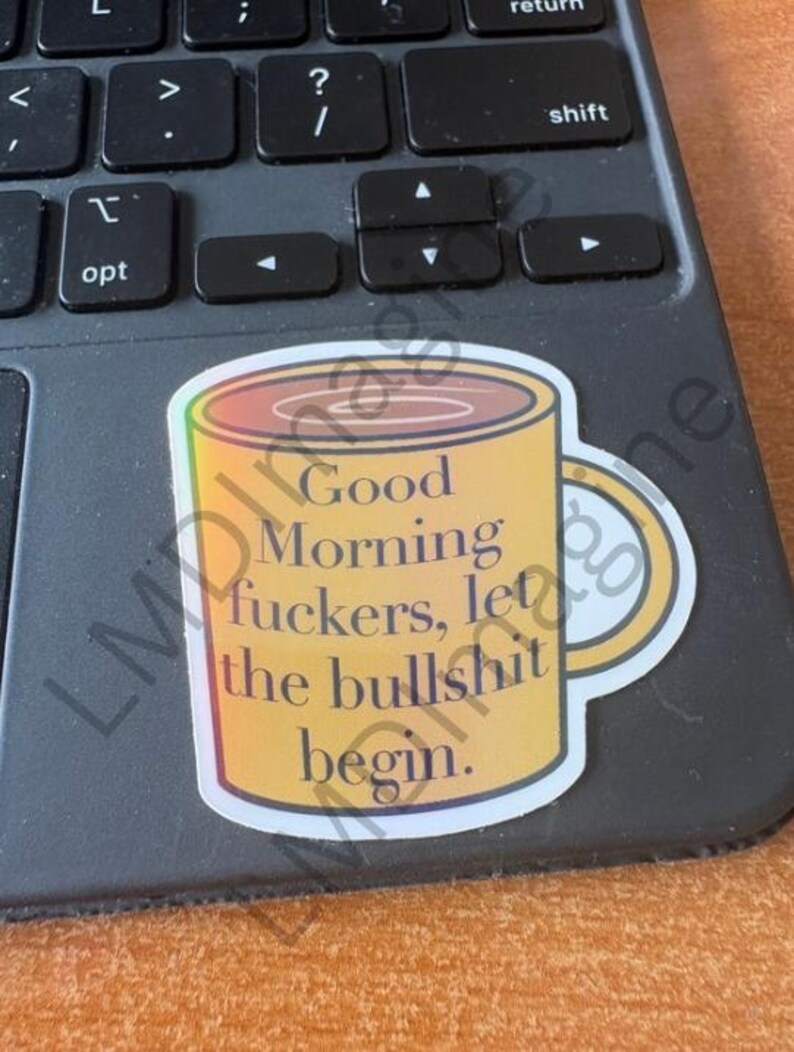 May include: A yellow coffee mug sticker with a gradient orange rim and handle. The text printed on the front of the mug reads, Good Morning fuckers, let the bullshit begin. The sticker is placed on a dark gray computer keyboard with black keys.