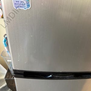 May include: A stainless steel mini refrigerator with a horizontal brushed metal finish and black trim. A small, die-cut sticker is adhered to the upper left corner, featuring a blue toaster illustration and the text FETCH ME MY BATHING TOASTER.