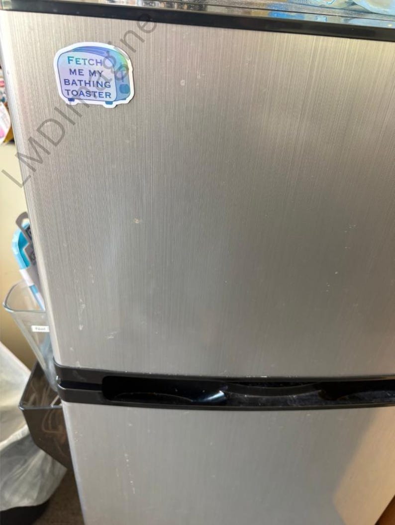 May include: A stainless steel mini refrigerator with a horizontal brushed metal finish and black trim. A small, die-cut sticker is adhered to the upper left corner, featuring a blue toaster illustration and the text FETCH ME MY BATHING TOASTER.