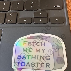 May include: A holographic sticker shaped like a toaster with the text FETCH ME MY BATHING TOASTER printed in black capital letters on the front. The sticker features a metallic, rainbow-reflective finish that shifts colors under light.