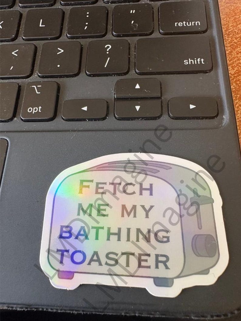 May include: A holographic sticker shaped like a toaster with the text FETCH ME MY BATHING TOASTER printed in black capital letters on the front. The sticker features a metallic, rainbow-reflective finish that shifts colors under light.