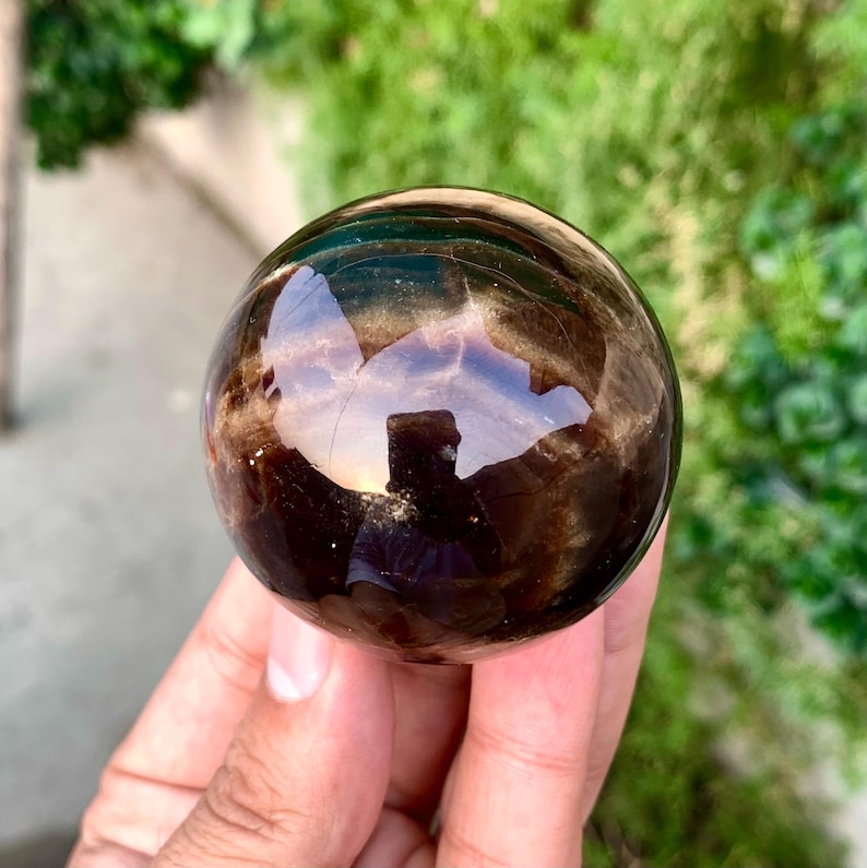 May include: A smooth, polished black obsidian sphere with iridescent rainbow flashes. The sphere is held in a hand.