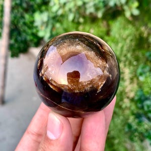 May include: A polished sphere of brown and black banded agate with a translucent center. The sphere is smooth and reflective, with a slight sheen.