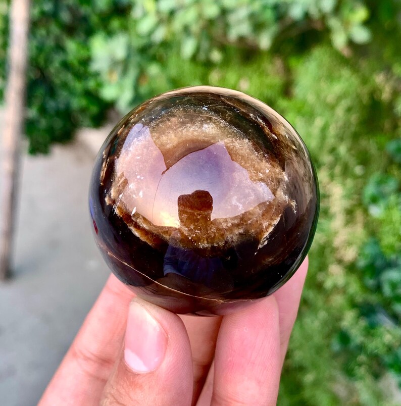 May include: A polished sphere of brown and black banded agate with a translucent center. The sphere is smooth and reflective, with a slight sheen.