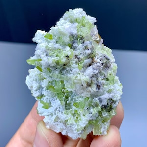 Puede incluir: Un grupo de cristales verde claro y blanco. Los cristales son de un color verde vibrante, con una mezcla de matriz blanca y gris. La forma general es irregular y texturizada, con una superficie rugosa. Los cristales están agrupados.
