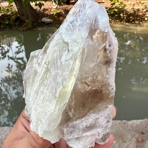 Könnte beinhalten: Ein rohes Kristallexemplar mit einer Kombination aus durchscheinenden blassgrünen und rauchbraunen Mineralformationen. Die Struktur weist gezackte, scharfe Kanten und eine geschichtete, kristalline Textur mit unterschiedlichem Klarheitsgrad auf.