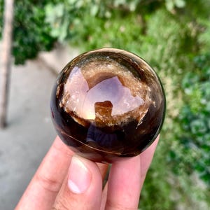 May include: A polished sphere of brown and black banded agate with a clear, translucent center. The sphere is smooth and reflective.