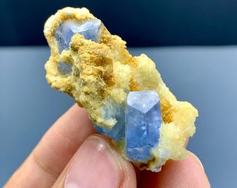 May include: A cluster of blue crystals with a hexagonal shape, set against a backdrop of tan and yellow mineral formations. The crystals are held in a hand, showcasing their natural beauty and geological structure.