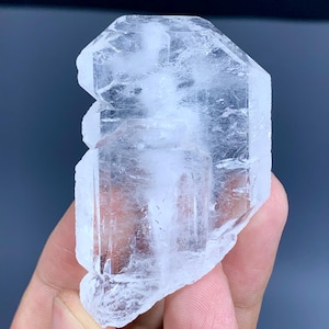 Puede incluir: Un espécimen mineral cristalino claro con una forma geométrica multifacética. La piedra transparente tiene una textura rugosa y natural y se sostiene en una mano. El interior del cristal muestra sutiles estructuras internas. El fondo es azul oscuro.