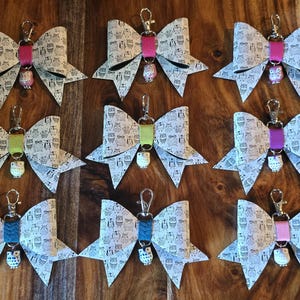 May include: Ten white bow keychains with owl charms. Each bow is made of fabric with a black and white owl print. The charms are different colors, including pink, green, blue, and teal.