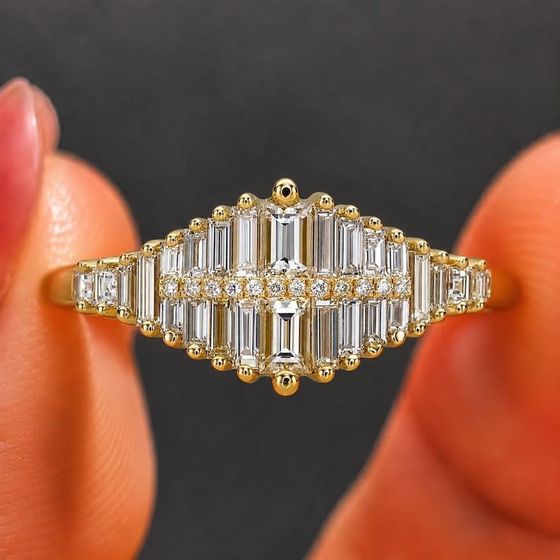 Close-up of a yellow gold diamond ring held between fingertips. The ring features a symmetrical geometric cluster of baguette-cut diamonds with a central vertical emerald-cut style stone, framed by round accent diamonds in an elongated Art Deco-inspired design.