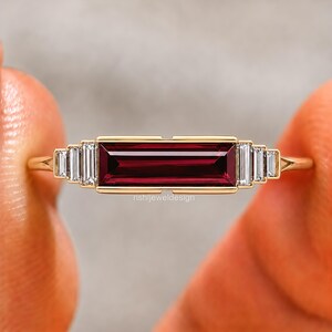 Op de afbeelding: Een gouden ring met een centrale horizontale baguette-geslepen dieprode granaatsteen, geflankeerd door drie kleinere baguette-geslepen witte diamanten aan elke kant. De tekst rishijeweldesign is zichtbaar op de zetting.