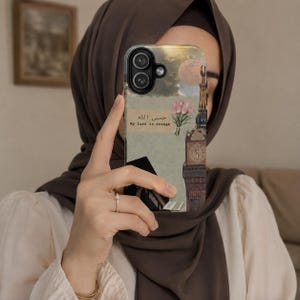 Peut inclure: Une coque de téléphone avec un design en collage représentant un ciel au clair de lune, un bouquet de tulipes roses, une tour d'horloge et un livre ouvert. Le texte "حسبي الله" et "My Lord is enough" est imprimé sur un fond beige papier.