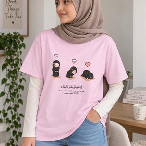 Könnte beinhalten: Ein hellrosa kurzärmeliges T-Shirt mit einer Grafik einer Zeichentrickfigur im Hidschab, die Gebetsschritte ausführt, mit rosa Herzen darüber. Der Text lautet: "And seek help through patience and prayer. [2:45]" sowie arabische Schrift.
