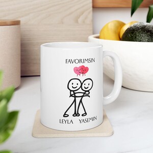 May include: White ceramic mug with a black and pink graphic. The graphic features two stick figures hugging, a pink heart, and the text "FAVORIMSIN", "LEYLA" and "YASEMIN". The mug is on a beige coaster.