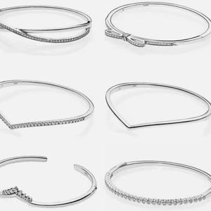 May include: Six silver-toned bangles with various designs. One features an overlapping design with embedded clear stones. Another has a bow-shaped accent with stones. Other bangles have V-shapes, open designs, and a row of stones.
