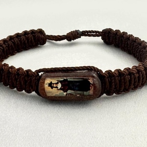May include: Brown woven bracelet featuring a rectangular wooden bead with a religious image. The bracelet is made of thick, braided cord and has an adjustable sliding knot closure. The central bead displays a detailed illustration.