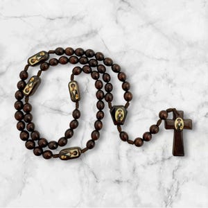 May include: A dark brown rosary with oval and rectangular beads. The rosary features a cross pendant and several rectangular beads with a small image of a person. The beads are connected by a dark brown string.