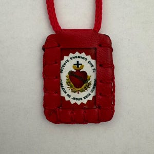 May include: Red leather pendant with a woven border and a red cord. The pendant features a central image of a heart with a cross and text in Spanish: "DETENTE ENEMIGO QUE EL CORAZON DE JESUS ESTA CONMIGO."