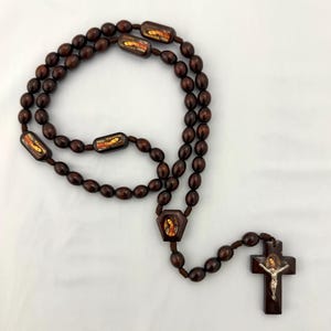May include: A dark brown rosary with oval beads and rectangular decorative pieces. The cross at the end features a depiction of Jesus. The rosary is made of wood and is laid out on a white surface.