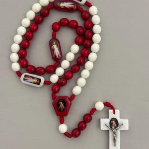 May include: A rosary with white and red beads. The rosary features a white cross with a silver Jesus figure, and red and white decorative pieces with religious imagery. The beads are strung on red cord.
