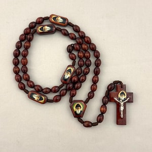 May include: A rosary featuring dark brown wooden beads strung on brown cord. The piece includes several rectangular and pentagonal wooden spacers, each displaying a small portrait of a bearded figure in a black habit. A wooden crucifix with a silver-toned metal corpus and a matching portrait of the same figure is attached at the end.