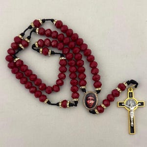 May include: A rosary featuring deep red faceted beads strung on a black cord, accented with small gold-tone spacers. The piece includes an oval pendant displaying a portrait of a face and a gold-tone crucifix with a silver-tone figure of Jesus.