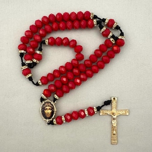 May include: A rosary featuring faceted bright red beads strung on black cord. Each bead is separated by a small gold-tone spacer set with clear crystals. The rosary includes an oval centerpiece depicting a face and a gold-tone crucifix with a figure.