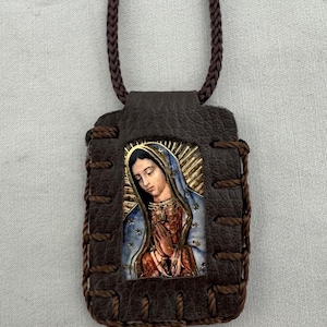 May include: A brown leather pendant featuring an image of the Virgin Mary, with a gold halo and blue and red robes. The pendant is stitched with brown thread and hangs from a braided brown cord.