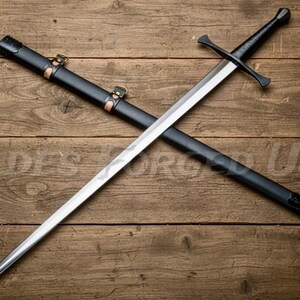 May include: A medieval sword with a silver blade and a black handle, resting on top of a black scabbard. The scabbard is secured with leather straps and bronze-colored hardware. The sword and scabbard are set against a wooden background.