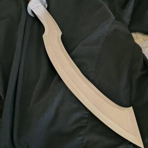 May include: A curved, tan-colored sword with a silver handle. The blade has a distinctive, crescent shape. The sword is lying on a dark, textured surface.