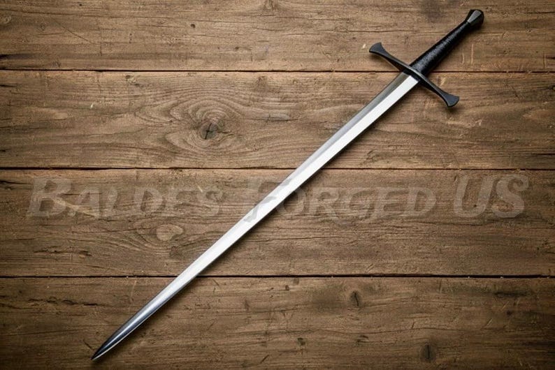 May include: A long, silver sword with a black handle and crossguard, resting on a wooden surface. The blade is sharp and pointed, and the handle has a rounded pommel. The text "BALDES FORGED US" is faintly visible on the wood.