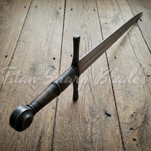 Peut inclure: Une &eacute;p&eacute;e de style m&eacute;di&eacute;val avec une longue lame &agrave; double tranchant. La poign&eacute;e et le pommeau sont en m&eacute;tal sombre, et la garde est de conception simple et droite. L'&eacute;p&eacute;e est pos&eacute;e sur une surface en bois.