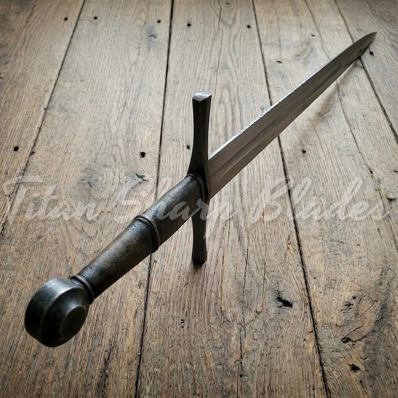 Peut inclure: Une &eacute;p&eacute;e de style m&eacute;di&eacute;val avec une longue lame &agrave; double tranchant. La poign&eacute;e et le pommeau sont en m&eacute;tal sombre, et la garde est de conception simple et droite. L'&eacute;p&eacute;e est pos&eacute;e sur une surface en bois.