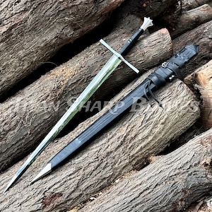 May include: A long, silver sword with a black handle and crossguard, resting on a pile of wooden logs. The sword is next to a black sheath with leather straps. The blade is shiny and reflects the light.
