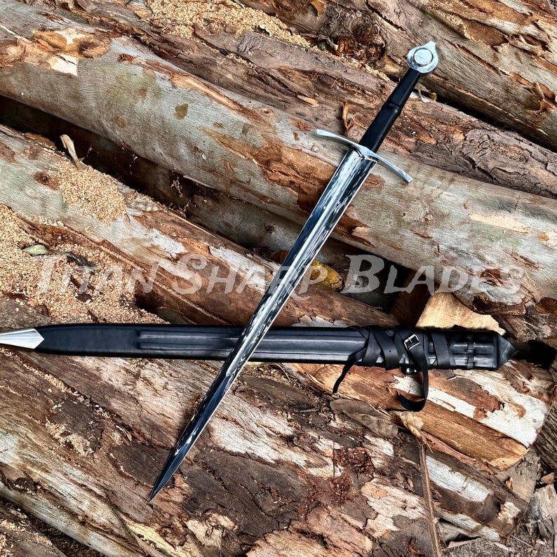 Pu&ograve; includere: Una spada in stile medievale con lama argentata e impugnatura nera, appoggiata su una superficie di legno. La spada &egrave; accompagnata da un fodero nero. Il testo "Iran Sharp Blades" &egrave; visibile.