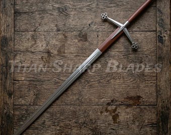 Peut inclure: Une longue épée argentée avec un manche et une garde transversale bruns. La garde transversale présente des motifs décoratifs en forme de trèfle. L'épée repose sur une surface en bois rustique. Le texte "Titan Sharp Blades" est visible.