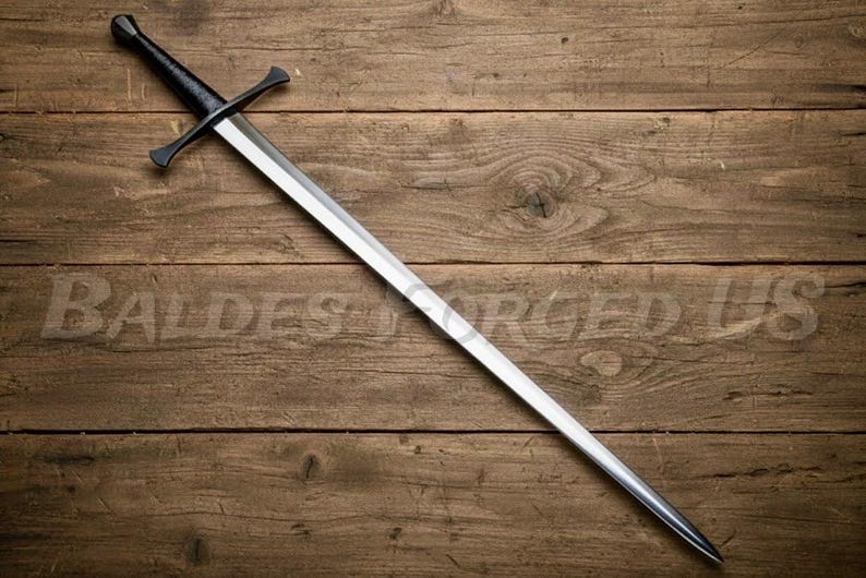 May include: A long, silver sword with a black handle and crossguard, resting on a wooden surface. The blade is polished and reflects light, while the handle and crossguard have a matte finish. The sword is a historical weapon.