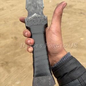 May include: A forged steel dagger with a textured blade and a wrapped black handle. The text "Blades Forged Us" is visible on the image. The dagger has a pointed tip and a wide, flat base.