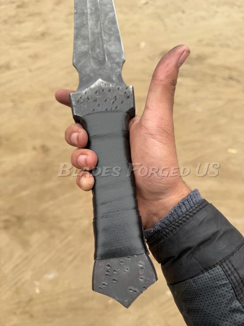 May include: A forged steel dagger with a textured blade and a wrapped black handle. The text "Blades Forged Us" is visible on the image. The dagger has a pointed tip and a wide, flat base.