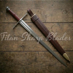 Può includere: Una spada in stile medievale con una lama argentata e un'impugnatura rivestita in pelle marrone. La spada è accompagnata da un fodero in pelle marrone con dettagli in ottone. Il testo "Titan Sharp Blades" è visibile.