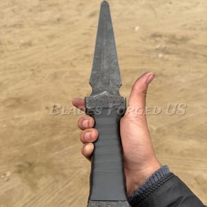 May include: A dark gray, hand-forged dagger with a long, pointed blade. The handle is wrapped in black material. The text "BLADES FORGED US" is visible on the image.