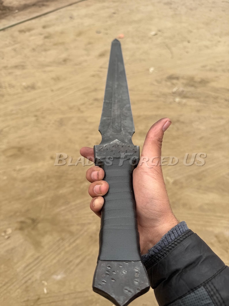 May include: A dark gray, hand-forged dagger with a long, pointed blade. The handle is wrapped in black material. The text "BLADES FORGED US" is visible on the image.