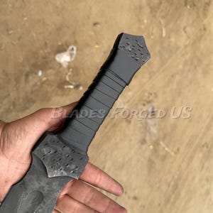 May include: A close-up of a black and gray sword with a textured handle and blade. The handle is wrapped with a dark material. The text "BLADES FORGED US" is visible.