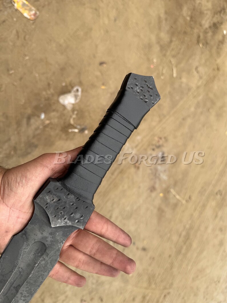 May include: A close-up of a black and gray sword with a textured handle and blade. The handle is wrapped with a dark material. The text "BLADES FORGED US" is visible.