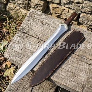 May include: A double-edged sword with a polished steel blade, a dark brown wooden handle with metallic rivets, and a matching dark brown leather sheath. The blade features a wide, leaf-shaped design that tapers to a sharp point at the tip.