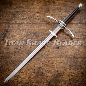 May include: A long, silver sword with a black handle and a cross-shaped guard. The blade is sharp and pointed, and the hilt features a decorative design. The text "Titan Sharp Blades" is visible on the wooden surface.