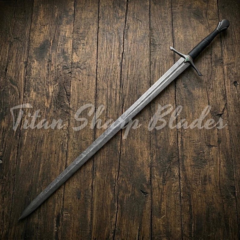 May include: A long, silver sword with a black handle and crossguard, resting on a wooden surface. The blade has a textured appearance. The text "Titan Sharp Blades" is visible on the wood.