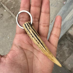 May include: A handmade keychain featuring a natural antler pendant with dark brown stripes. The pendant is attached to a silver-colored metal keyring. The keychain is held in a hand.