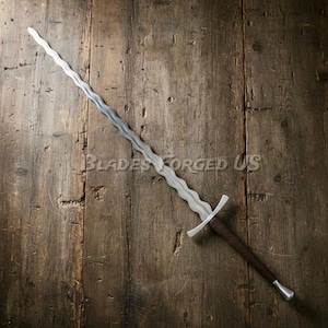 Op de afbeelding: Een lang zwaard met een golvend lemmet, bruin handvat en zilveren pommel. Het lemmet heeft een uniek, golvend ontwerp. De tekst "Blades Forged US" is zichtbaar op het houten oppervlak. Het zwaard ligt op een rustieke houten achtergrond.